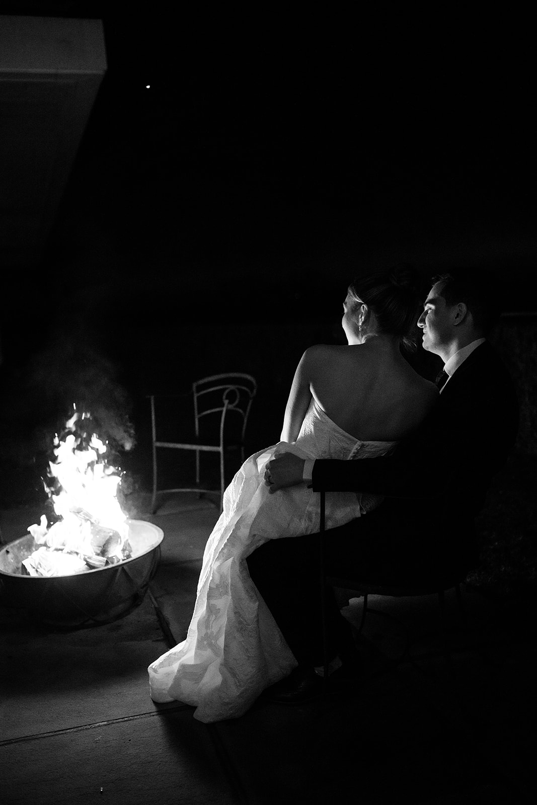 Black and white night photo from behind of the couple sitting together and watching a warm bonfire fire pit.