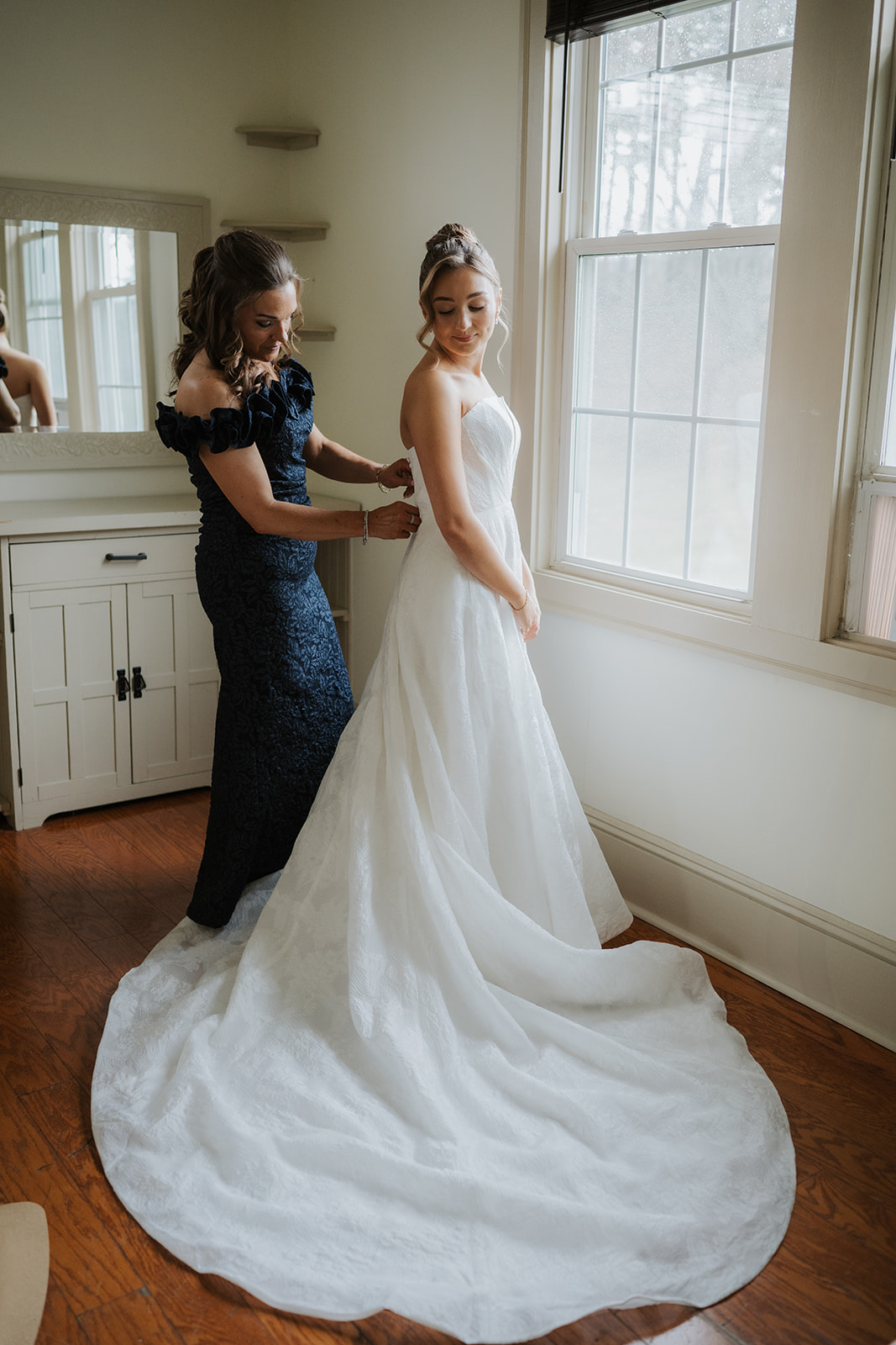 Wider shot of the mother buttoning the bride's gown as they stand in front of the window, showing the full train.