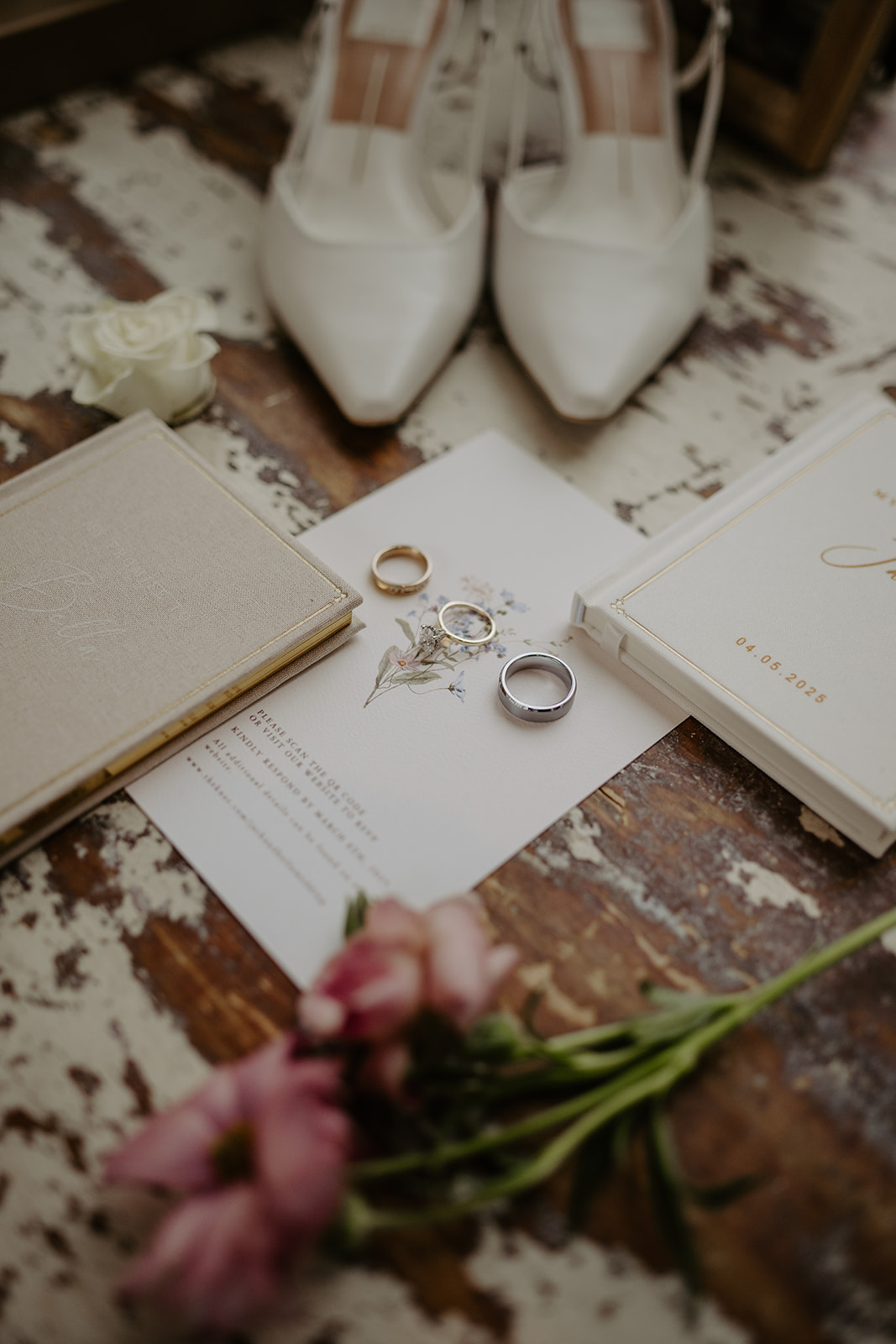 Flatlay of the wedding invitation card, vows booklets, wedding rings, white bridal heels, and flowers on a textured table.