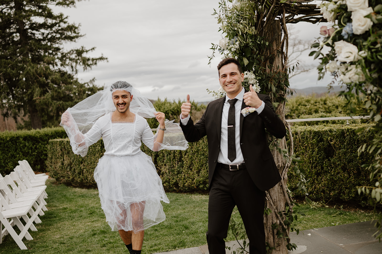 Groom smiling and giving two thumbs up to the camera, while the best man in the tutu/veil walks up laughing in the background.