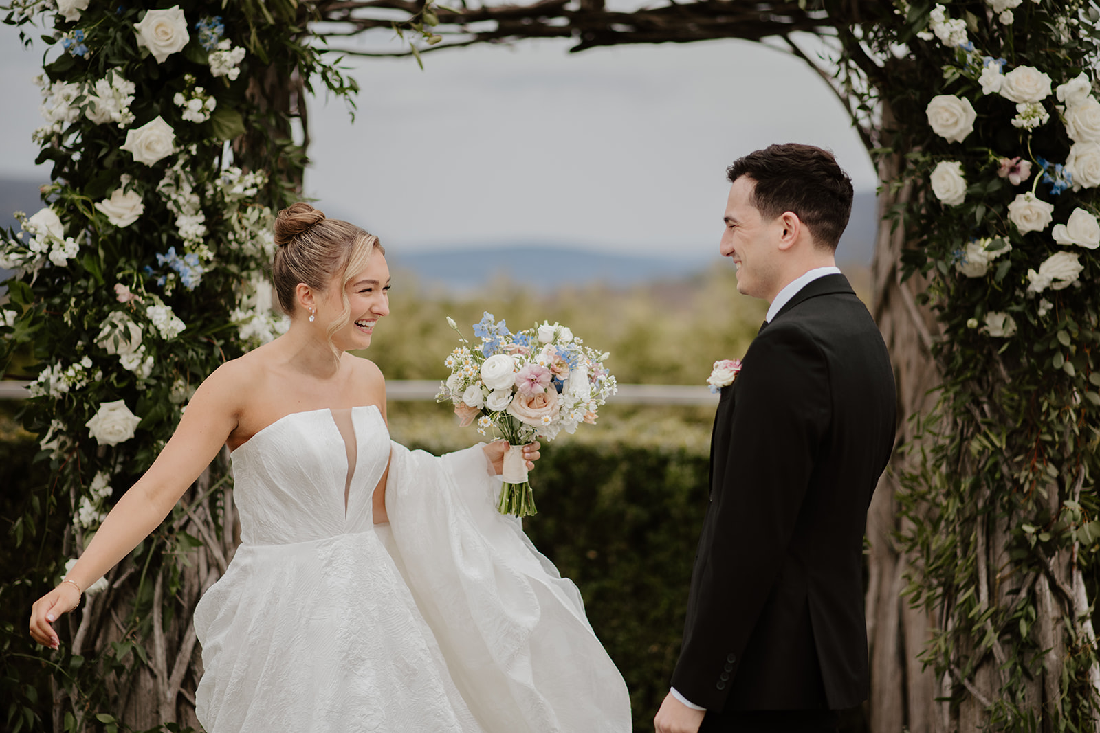 Bride and groom looking at each other and laughing happily after he turned around, standing under the wooden arch decorated with white roses at The Garrison in Garrison, NY.