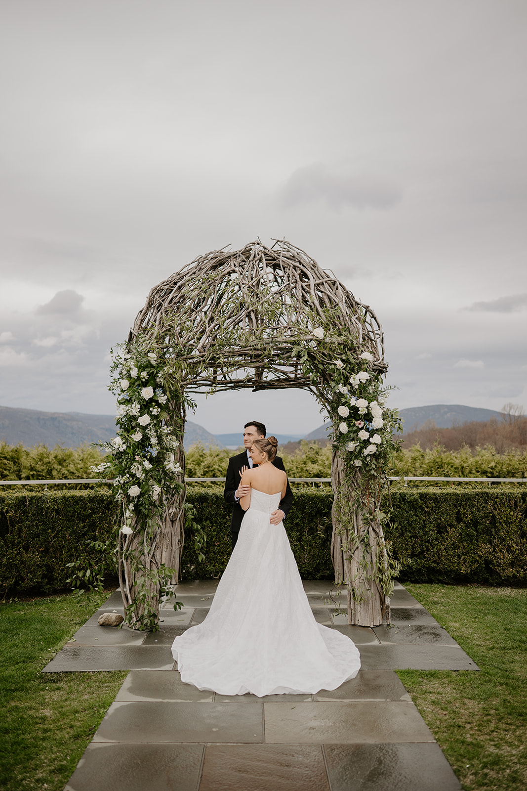 Couple under the arch, back view of the bride showing the full train of her wedding gown, looking away towards the side.