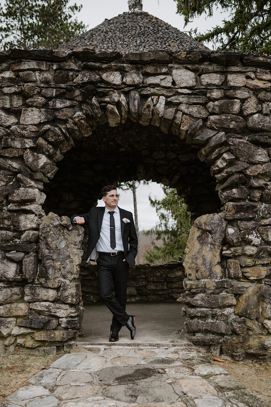 Groom leaning against a round stone gazebo wall, looking thoughtful.