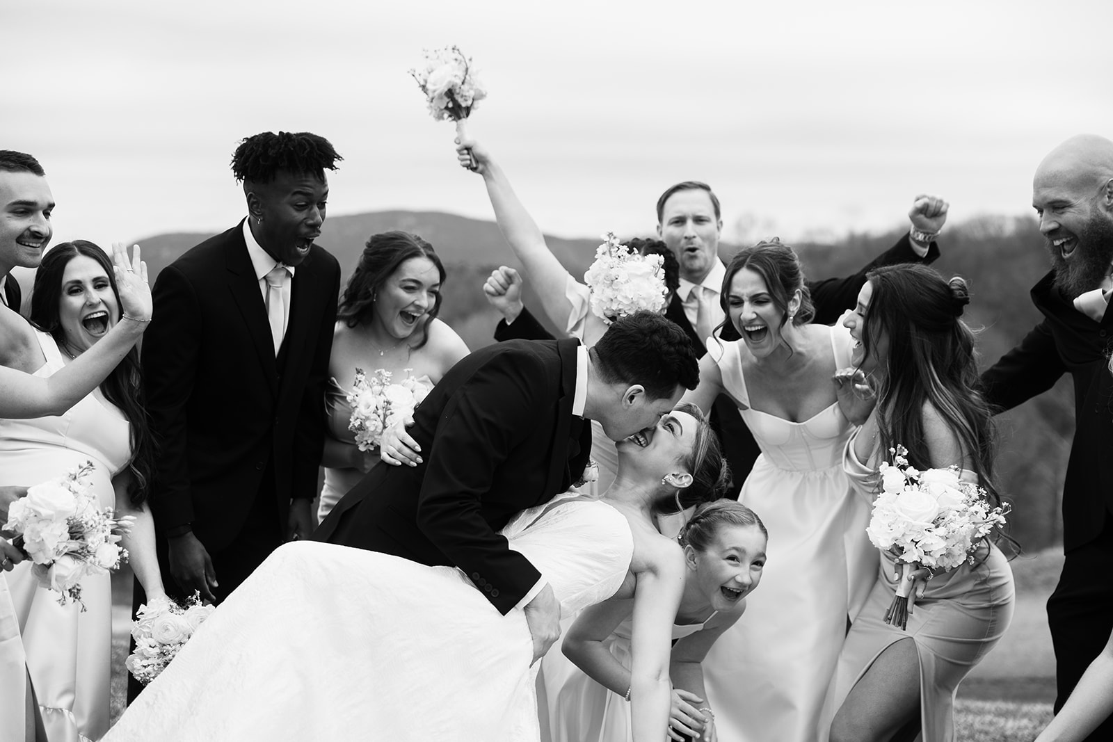 Black and white photo of the wedding party cheering as the groom dips and kisses the bride.