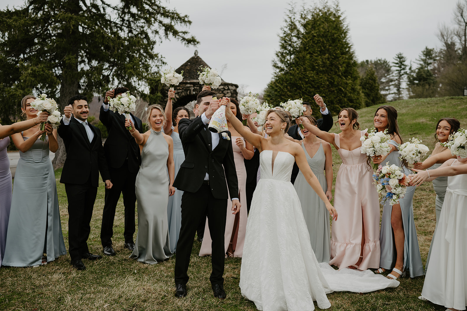Bridal party cheering on the lawn as the groom prepares to pop a champagne bottle with the bride.