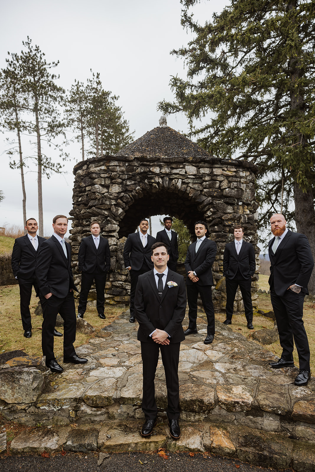 Groom standing in front of the stone gazebo with the groomsmen in a V-formation behind him.