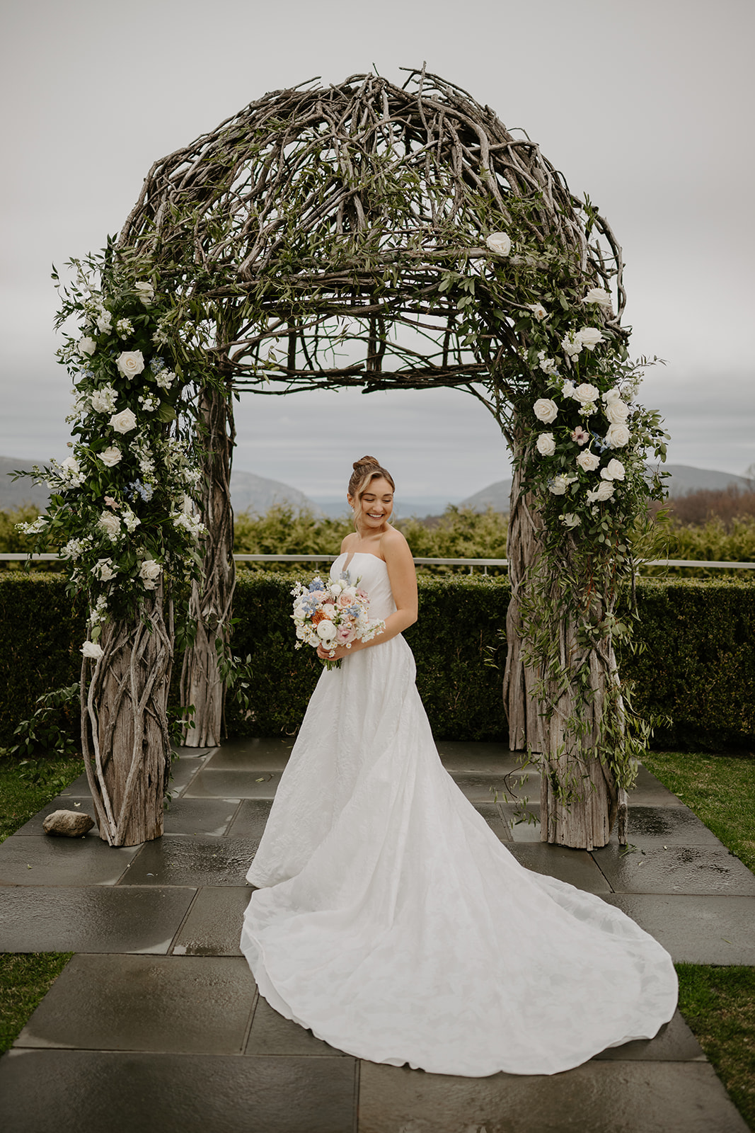 Bride standing under the ceremony arch, holding her bouquet and looking down.