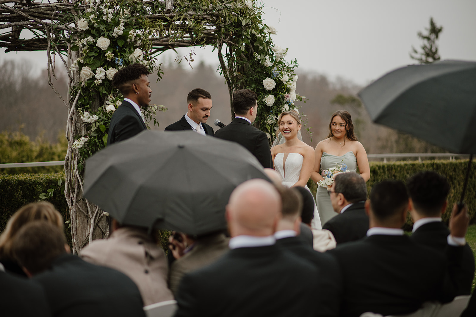 Bride and groom laughing at the altar while the officiant reads vows, guests holding clear umbrellas in the foreground.