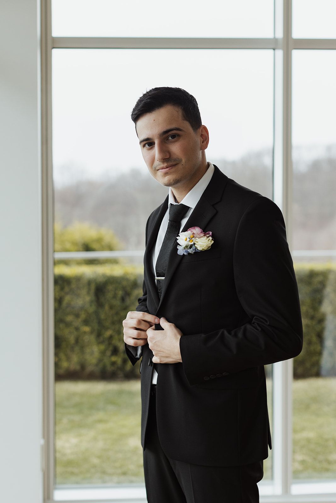 Groom buttoning his tuxedo jacket, looking at the camera, with a boutonniere on his lapel.