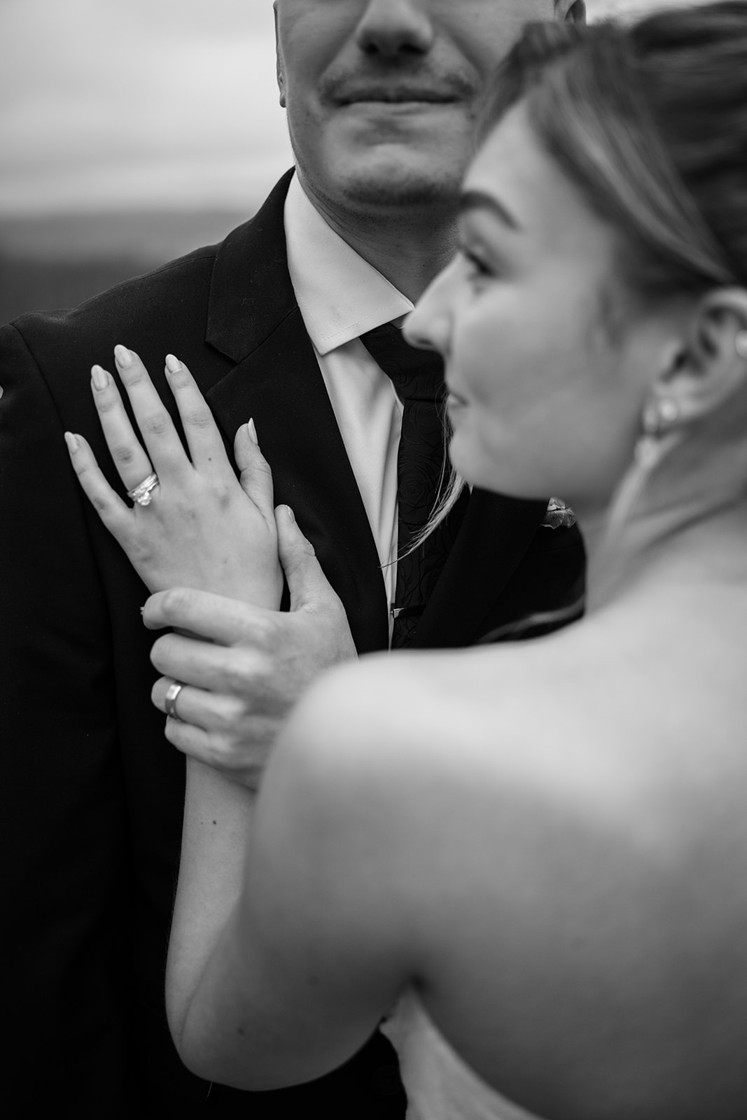 Close-up black and white photo of the bride's hand with her engagement and wedding rings resting on the groom's tuxedo jacket.