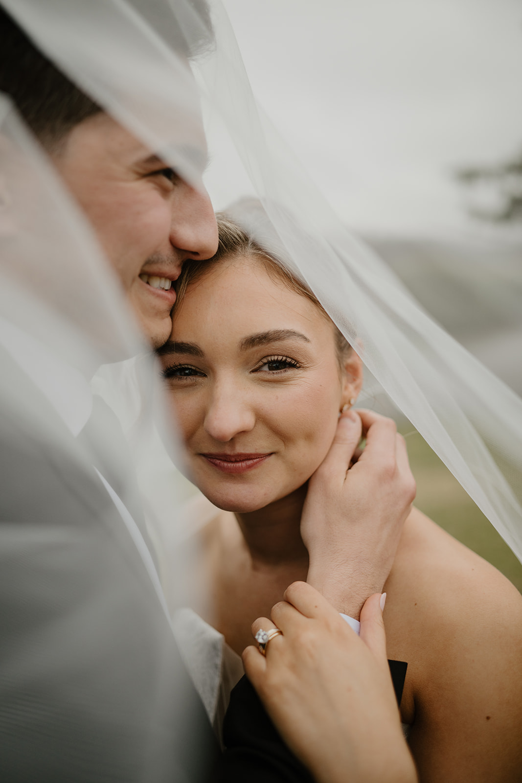Color close-up of the bride smiling softly under her veil while the groom gently touches her face.