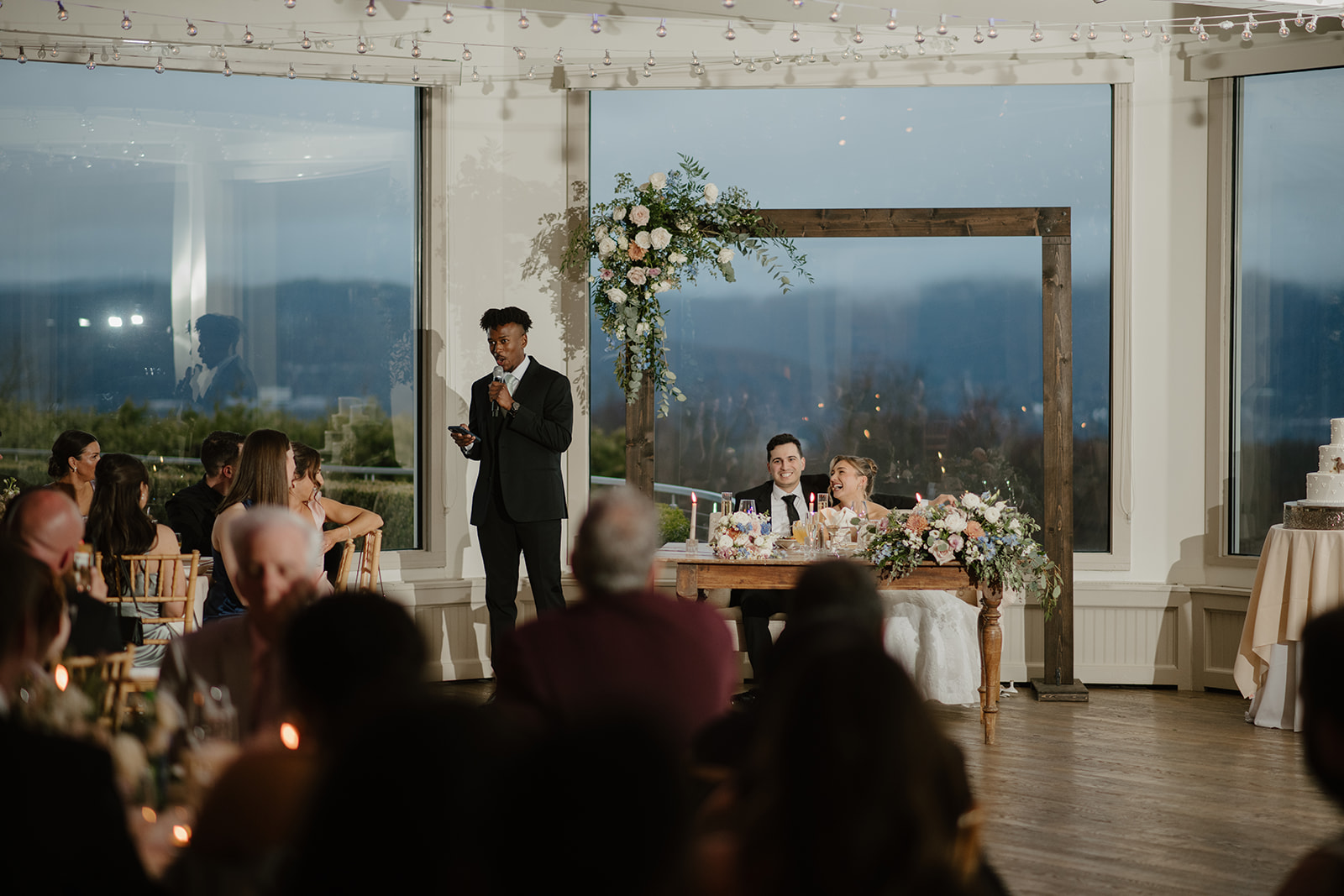 Best man giving a speech with a microphone, while the newlyweds listen from their sweetheart table decorated with floral arrangements.