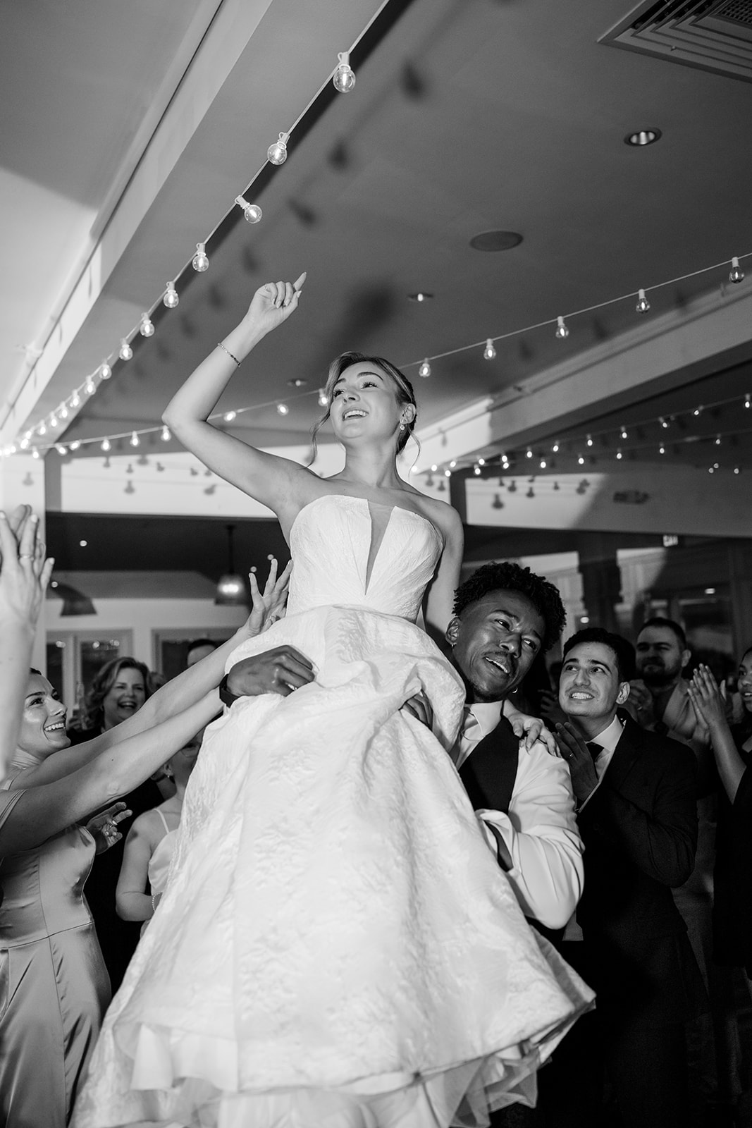 Black and white photo of the bride laughing and pointing to the sky while lifted on shoulders during the wedding reception party.