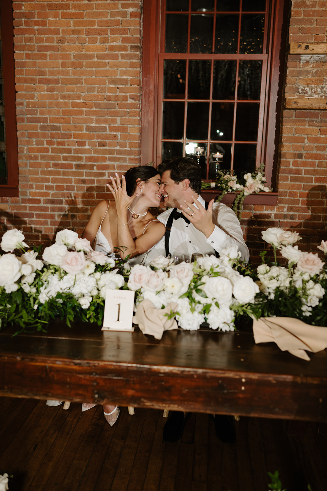 Bride and groom smile happily at their sweetheart table while showing off their wedding bands.