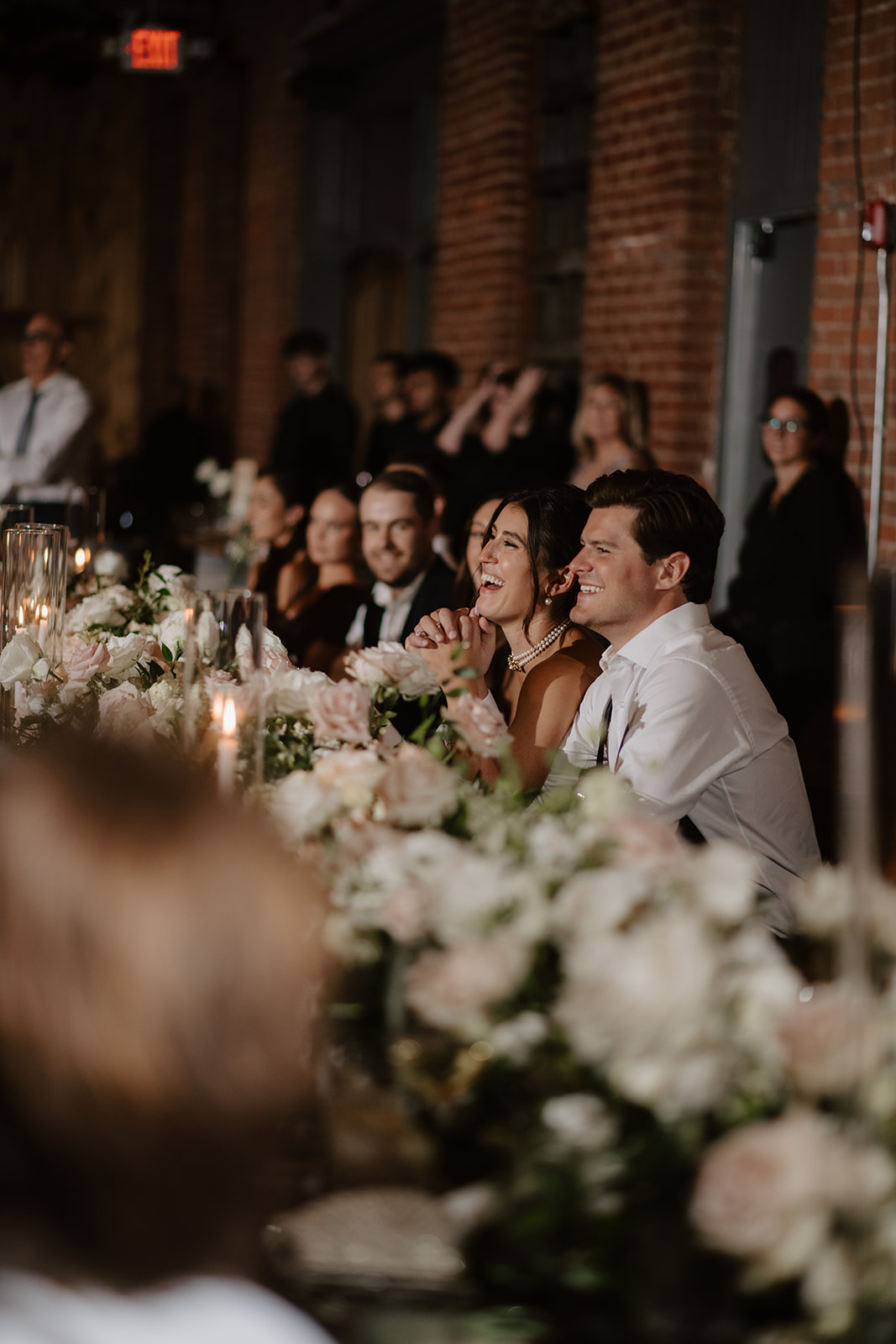 Bride and groom laugh heartily while listening to speeches at their flower-adorned head table.