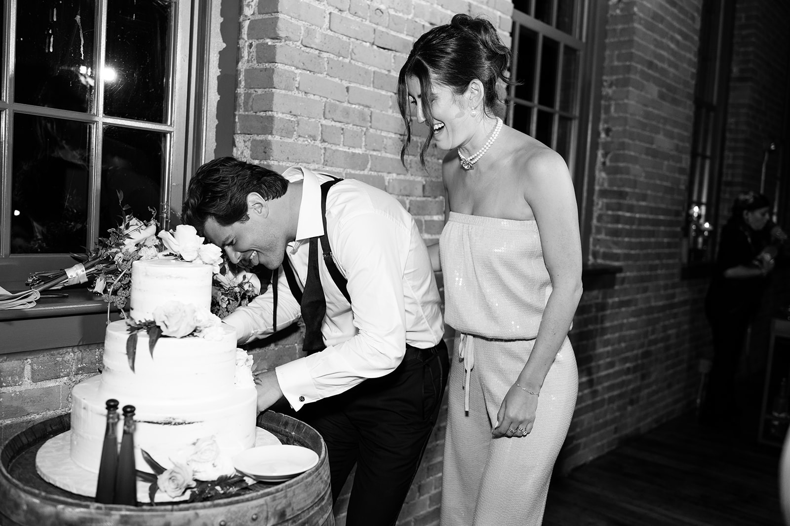 Black and white portrait of the bride laughing as the groom leans in to bite the wedding cake.