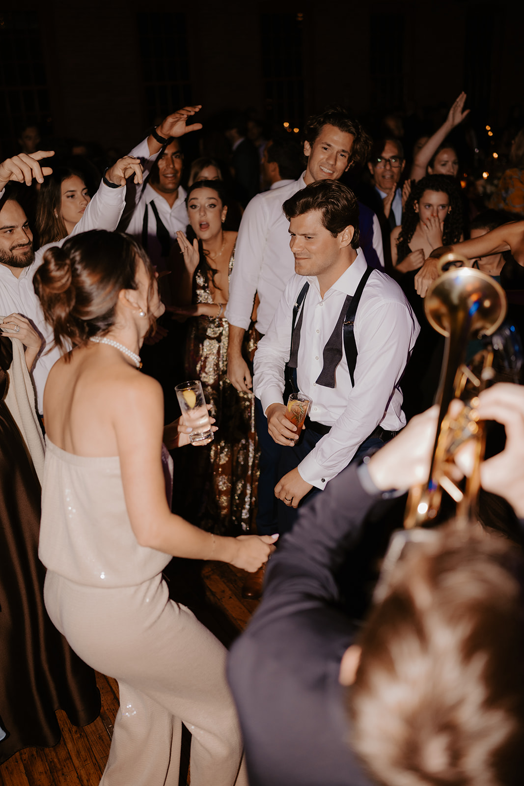 Bride and groom dance with cocktails in hand on a packed reception dance floor with a trumpet player.