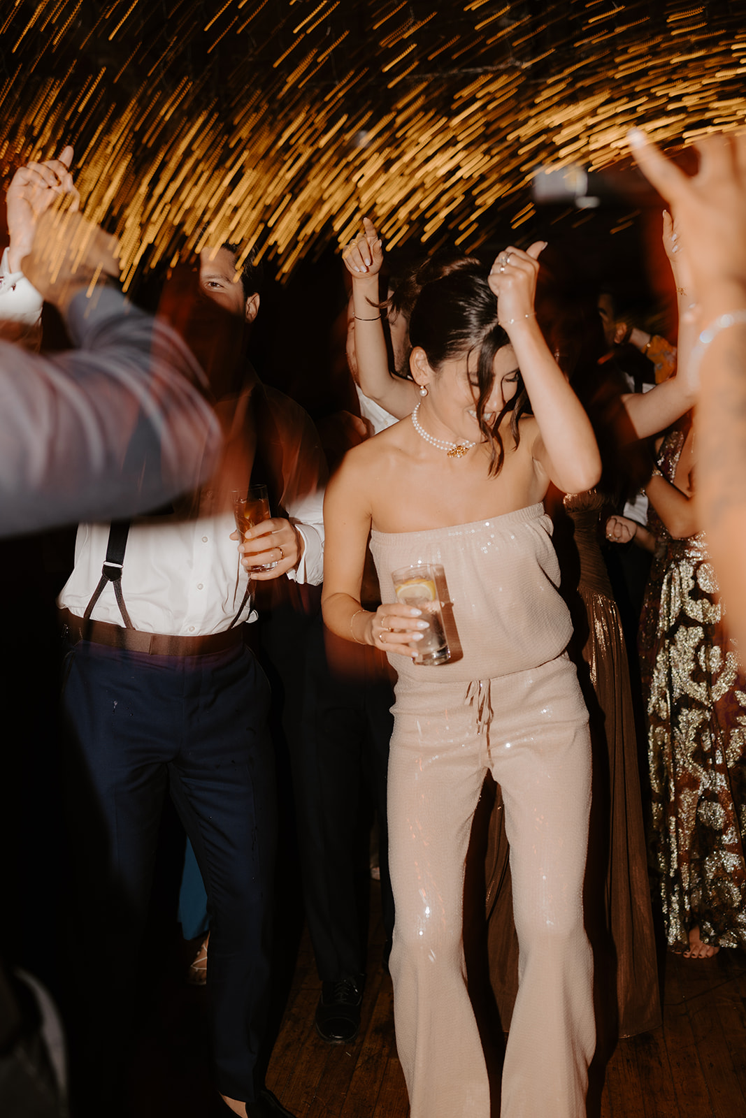 Bride in a sparkly jumpsuit dances joyfully under the warm reception lights.