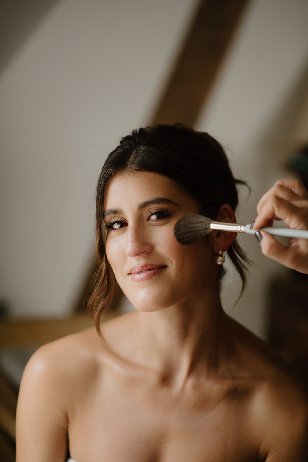 Bride smiles gently as makeup is applied to her cheek with a soft brush during preparations.