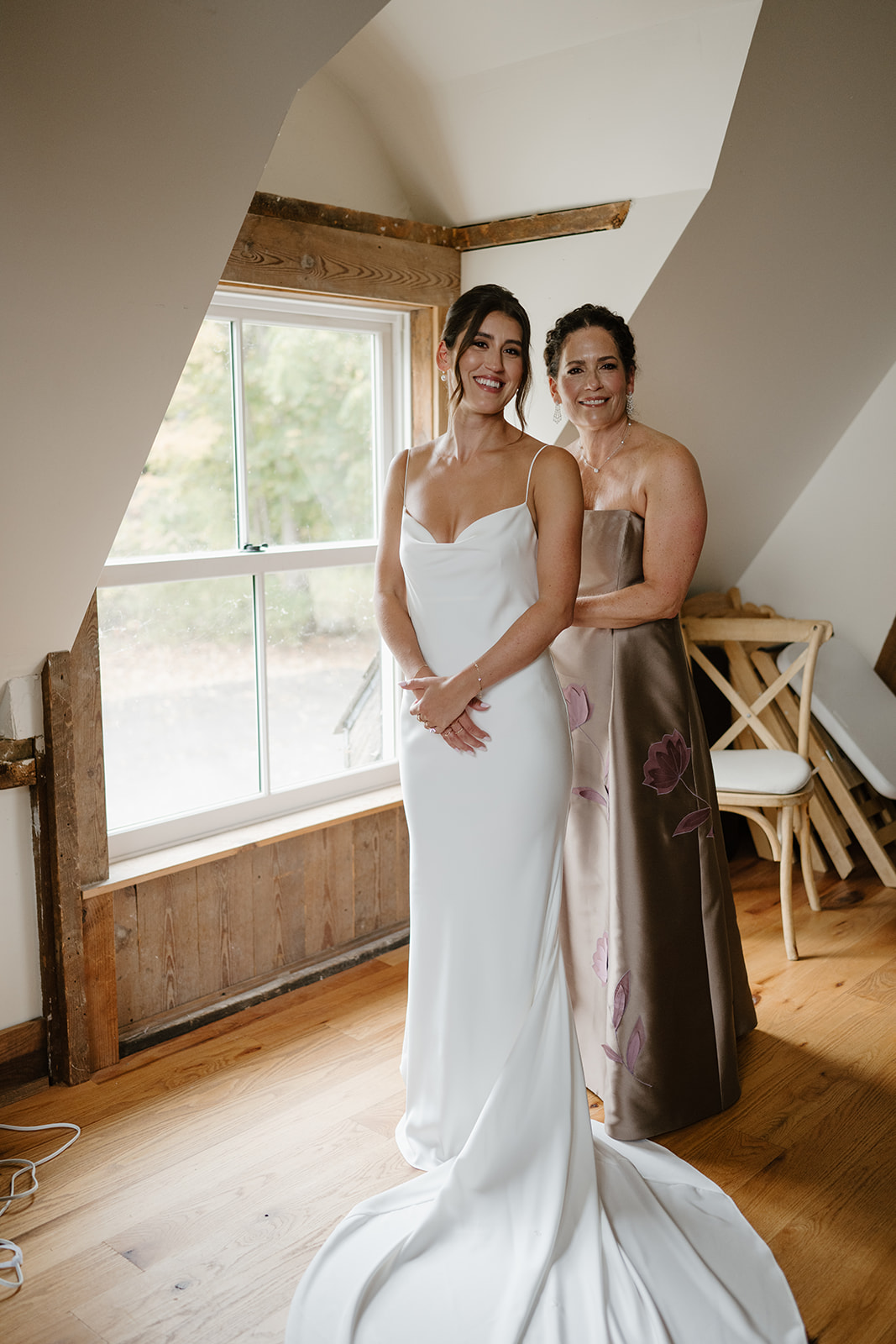Bride and her mother smile for a portrait in a rustic, sunlit dressing room.