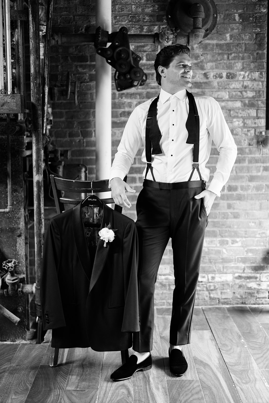 Black and white photo of the groom posing in suspenders next to his wedding jacket on a chair.