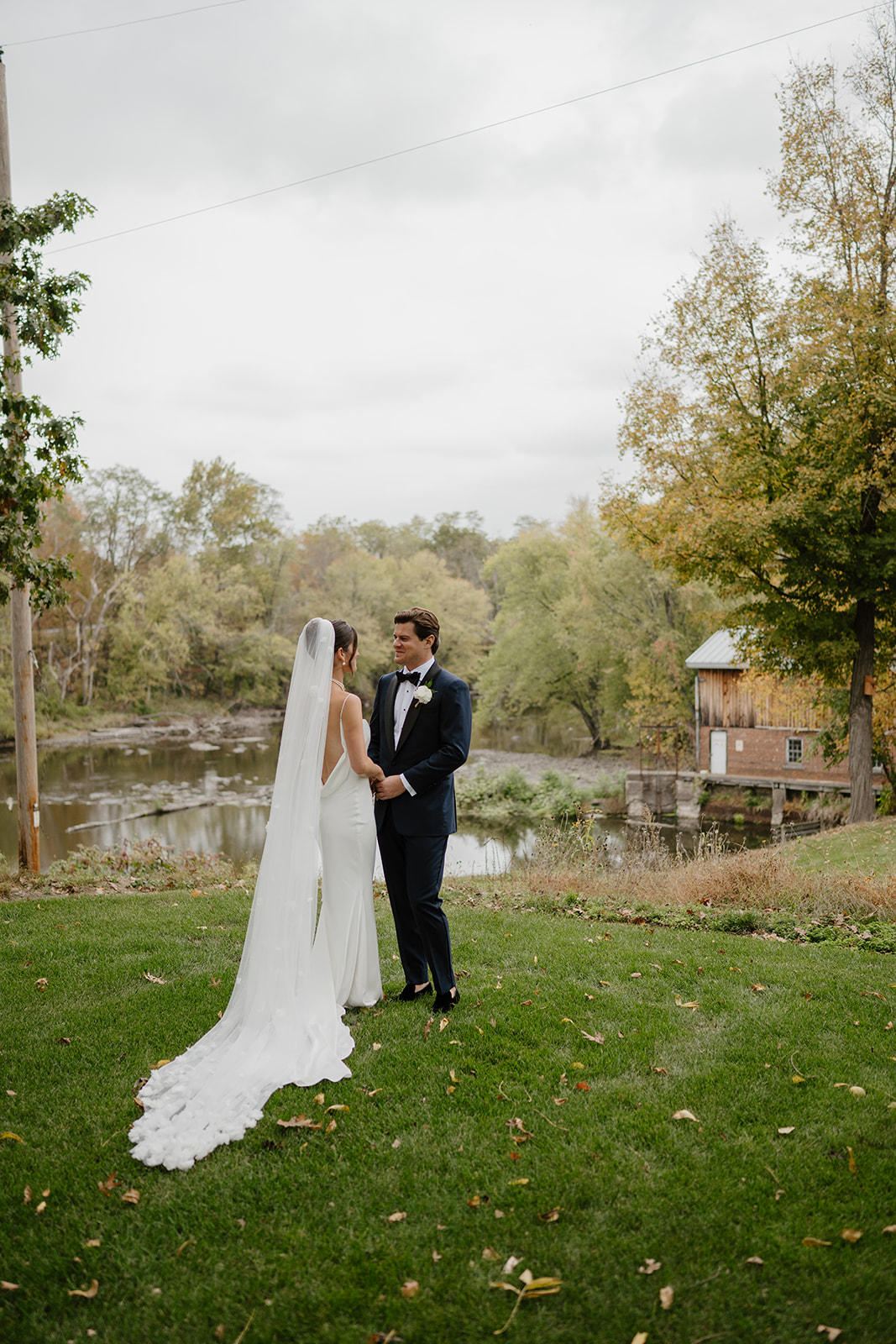 Bride and groom hold hands by the riverbank with historic buildings and trees in the background.