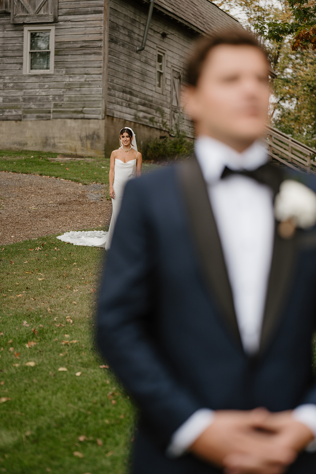 Bride stands in focus in front of a rustic barn as the groom waits out of focus in the foreground.