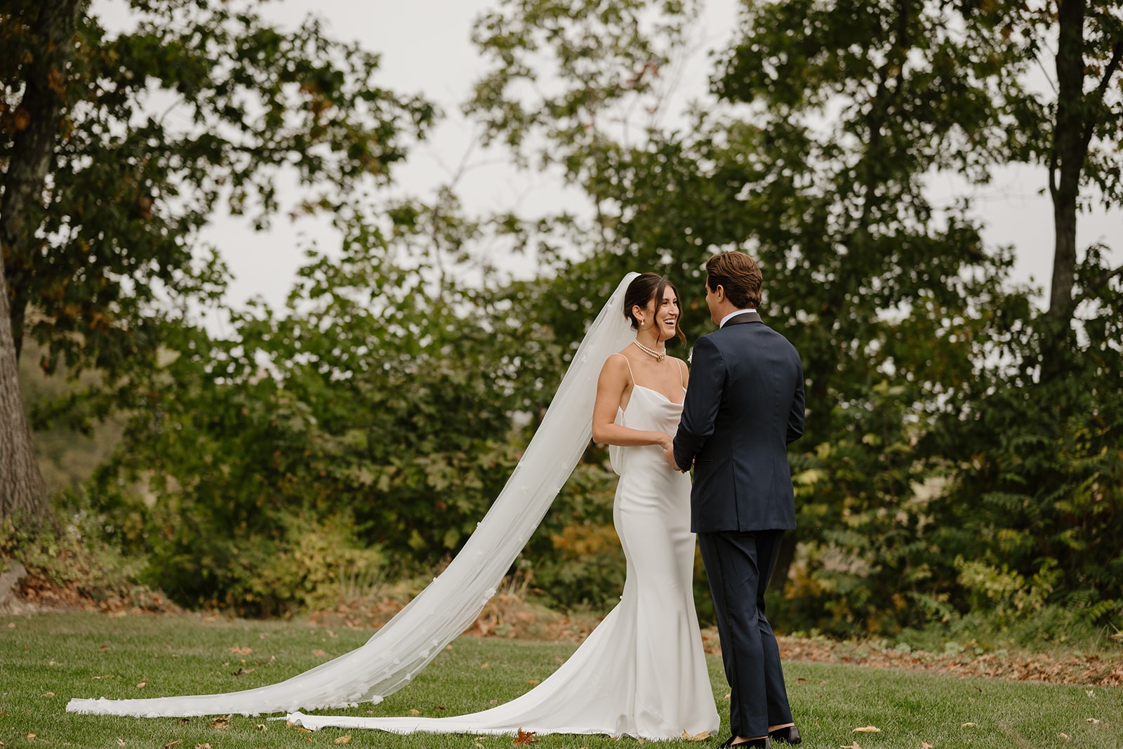 Bride and groom stand smiling on a green lawn, the bride's long tulle veil draping behind her.