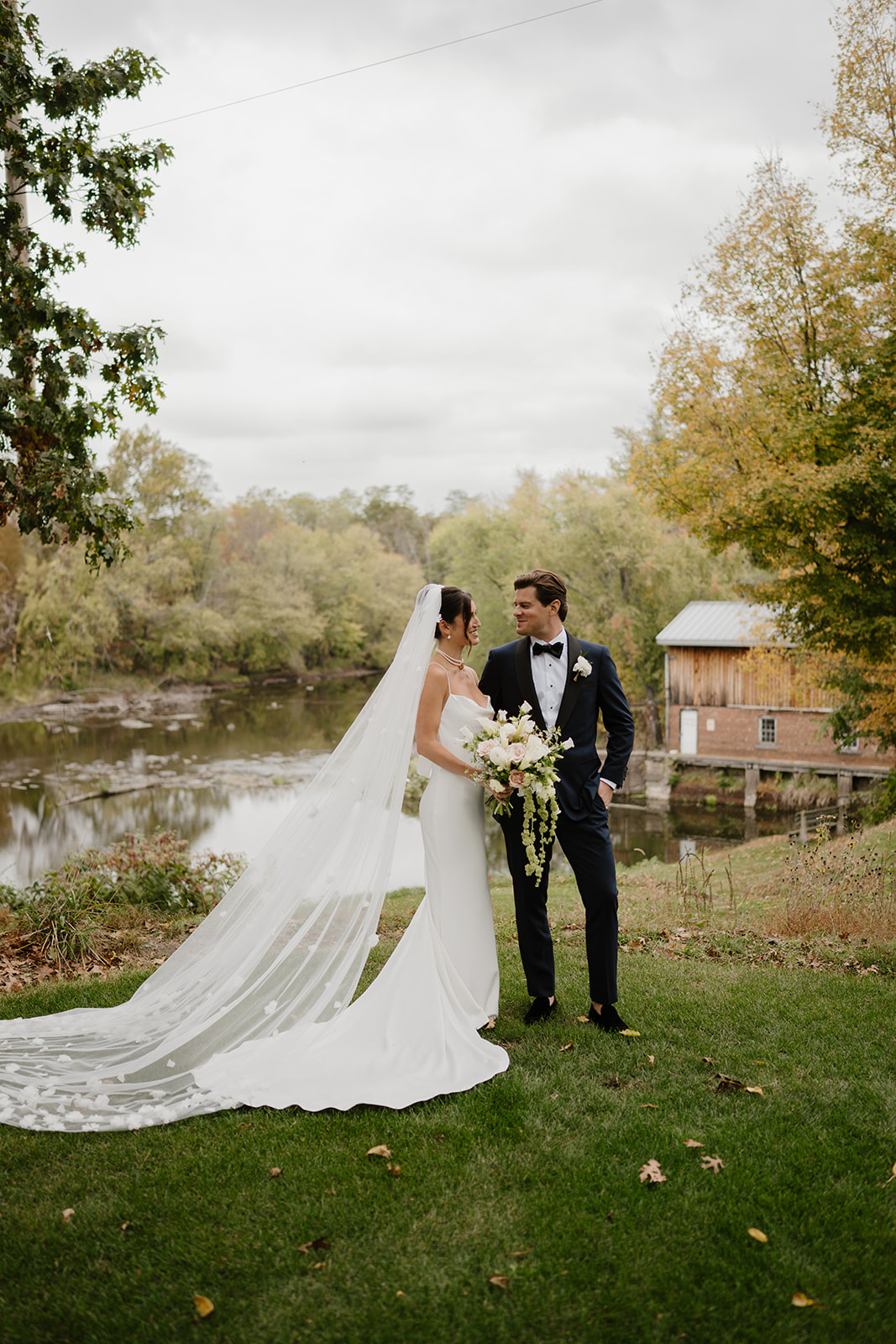 Couple smiles at each other by a river, the bride holding a white floral bouquet with a long veil.