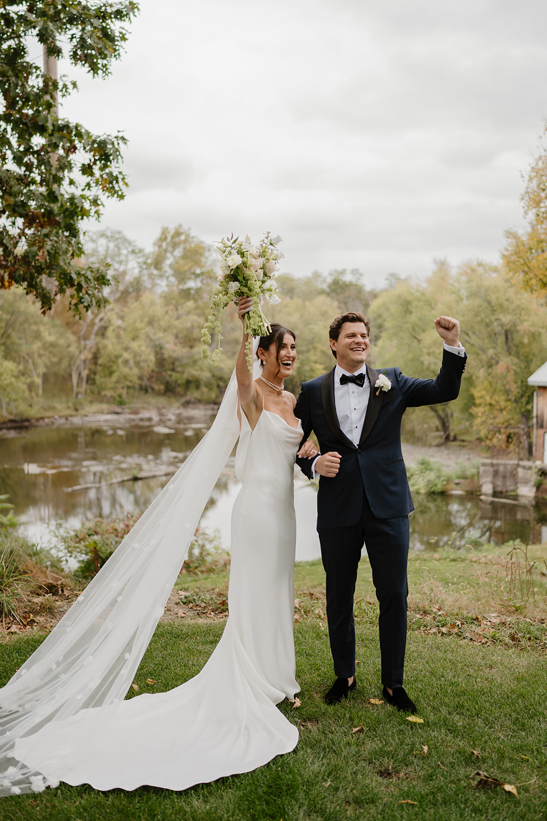 Bride and groom cheer with raised bouquet and fist pump by the scenic riverbank.