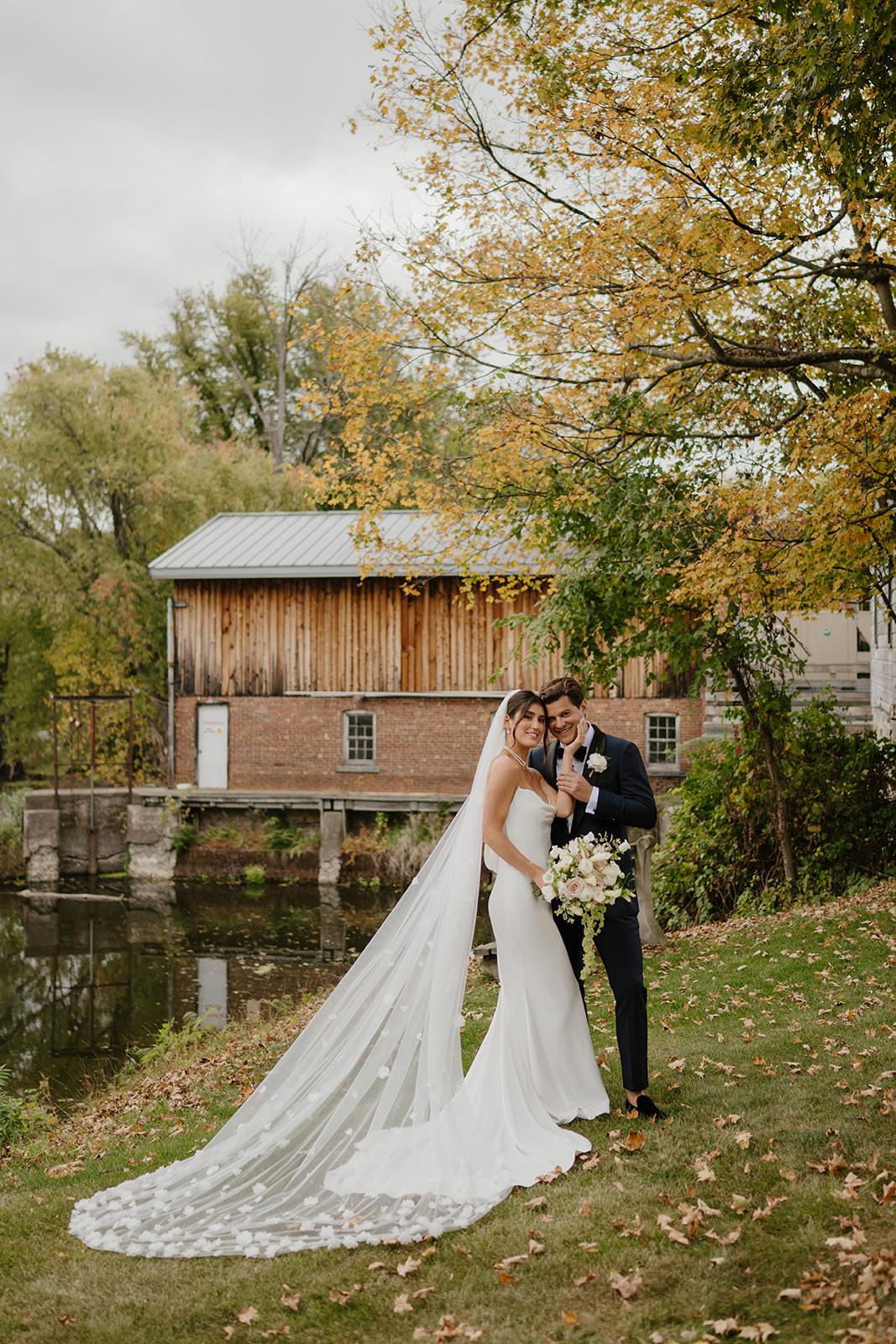 Bride and groom embrace by a calm river with a historic brick mill in the background.