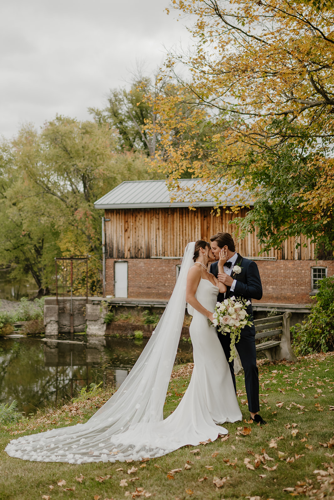 Bride and groom share a romantic kiss by a calm river in front of a rustic mill.