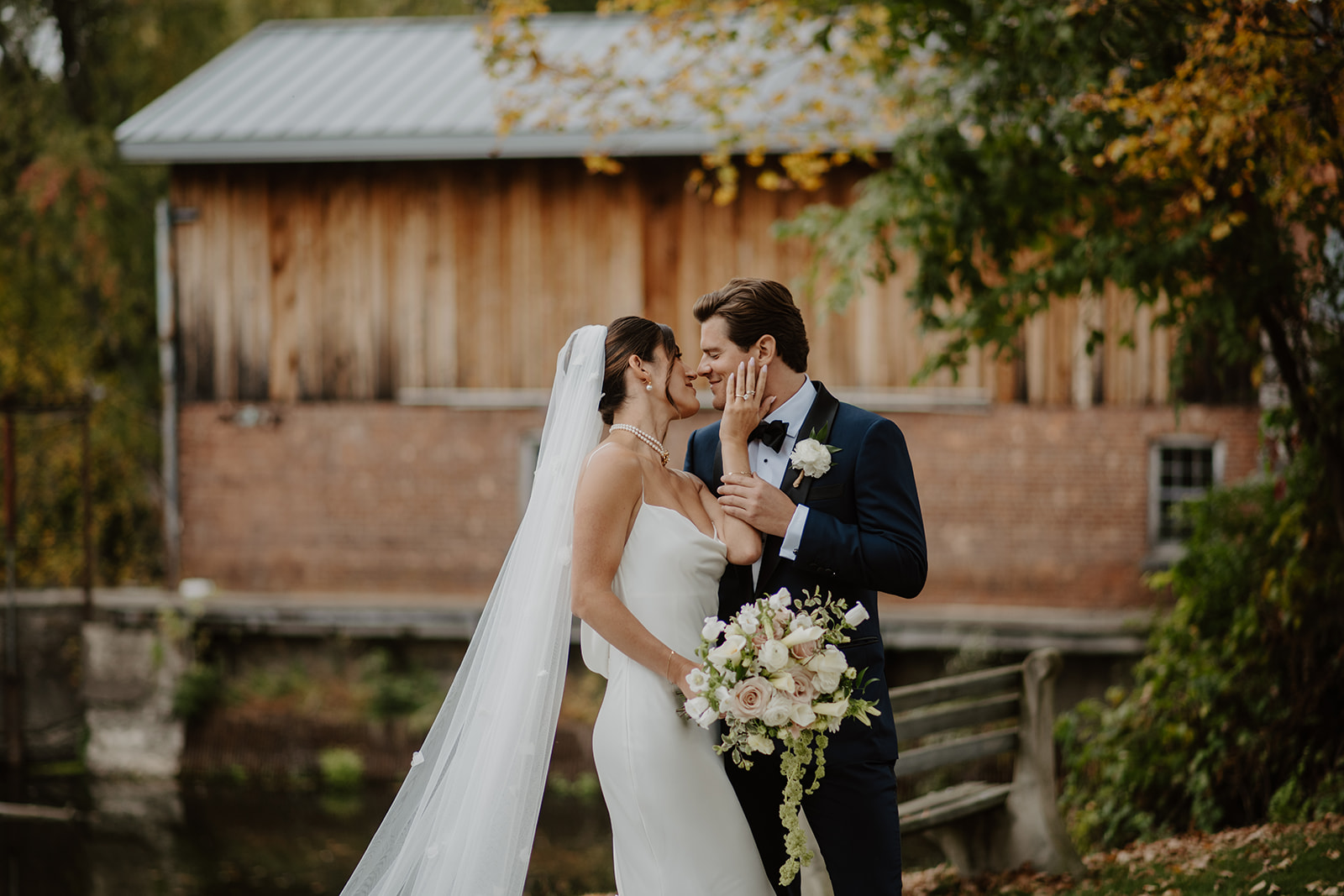 Bride gently touches the groom's face as they share a sweet moment in front of a rustic mill.