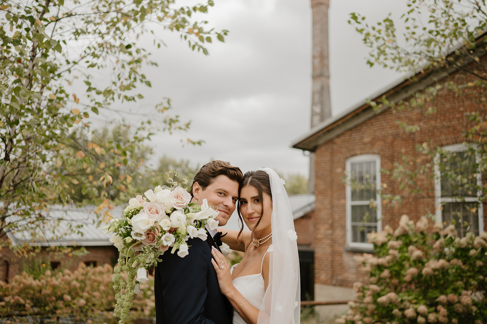 Bride and groom look tenderly at the camera, posing with a lush white floral bouquet.