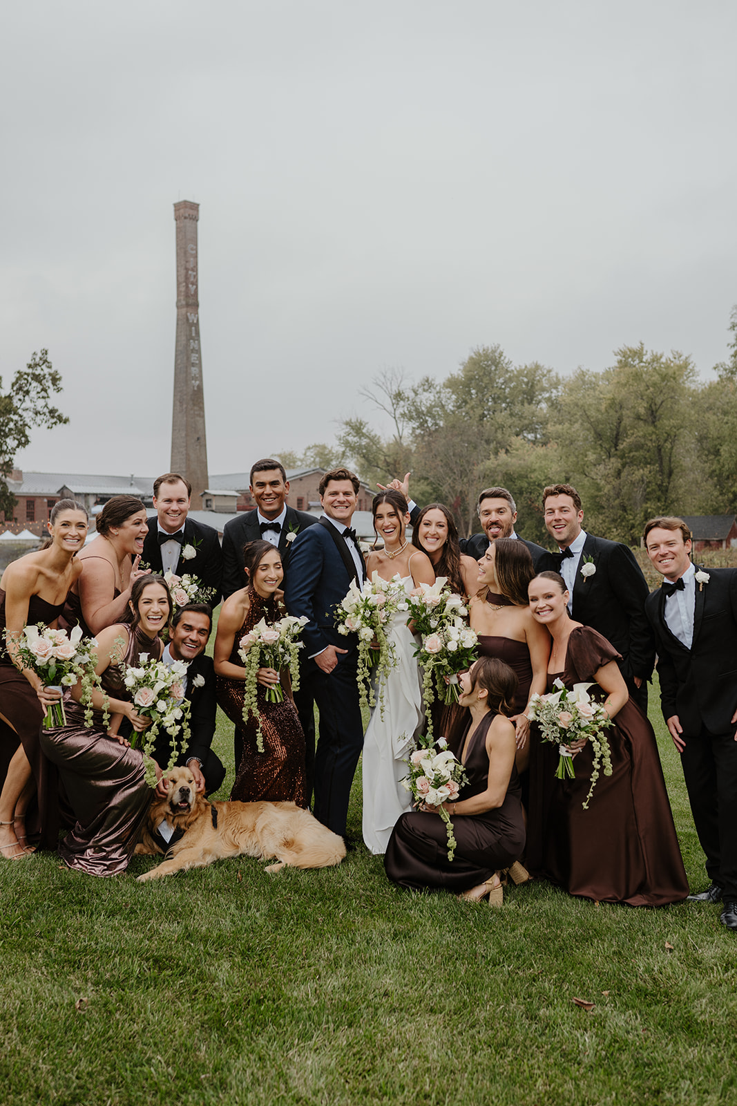 Wedding party in brown dresses and navy tuxedos poses with their golden retriever on the lawn.