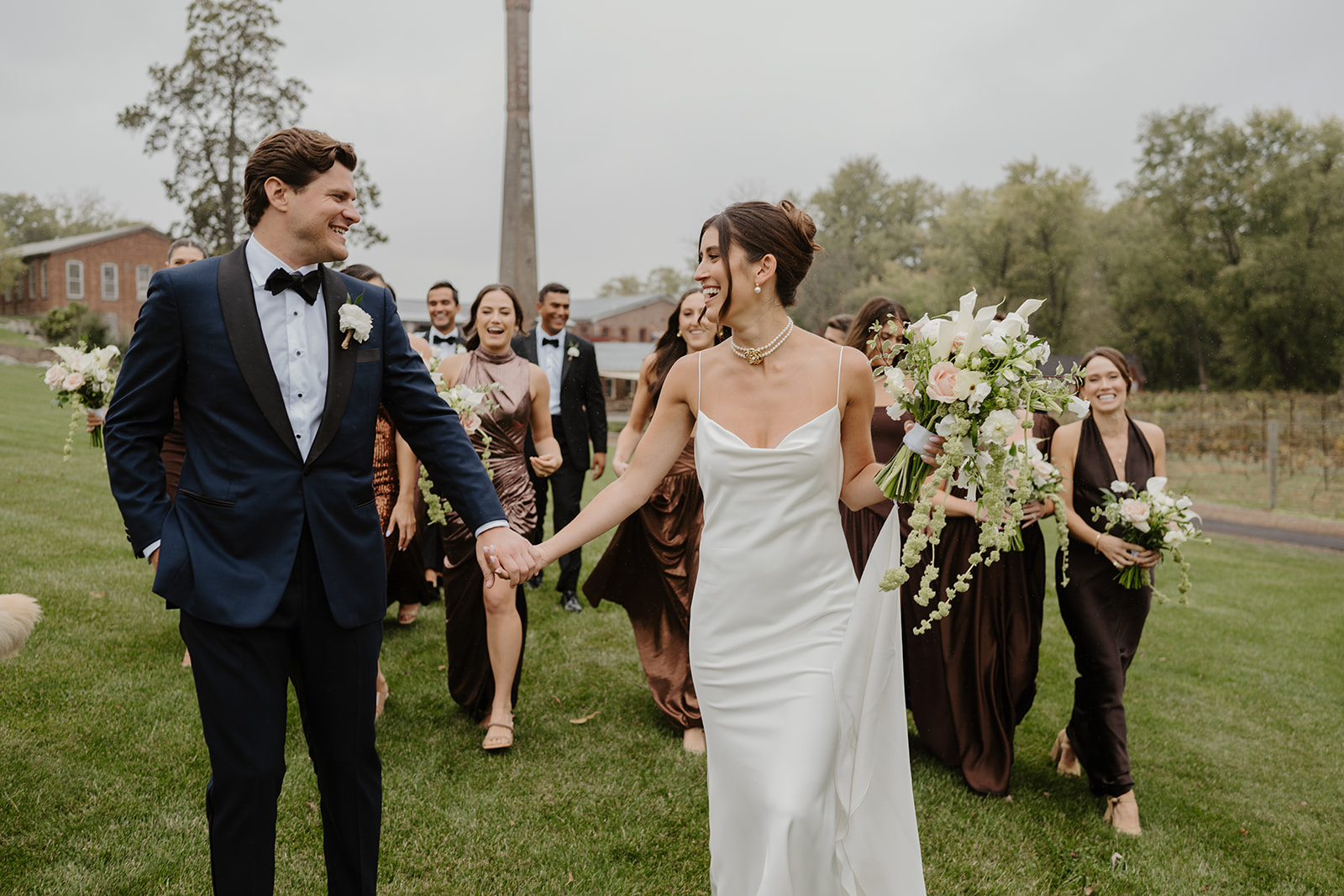 Bride and groom lead their wedding party across the lawn while smiling and holding hands.