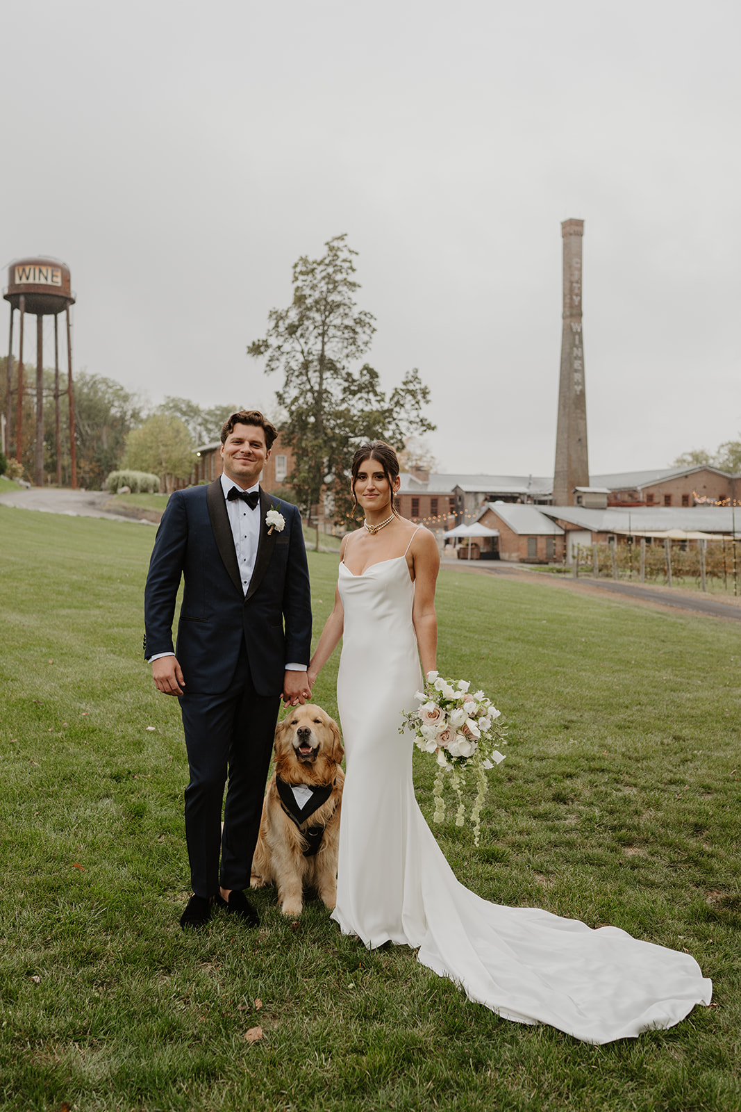 Bride and groom stand hand-in-hand with their golden retriever on the City Winery lawn.