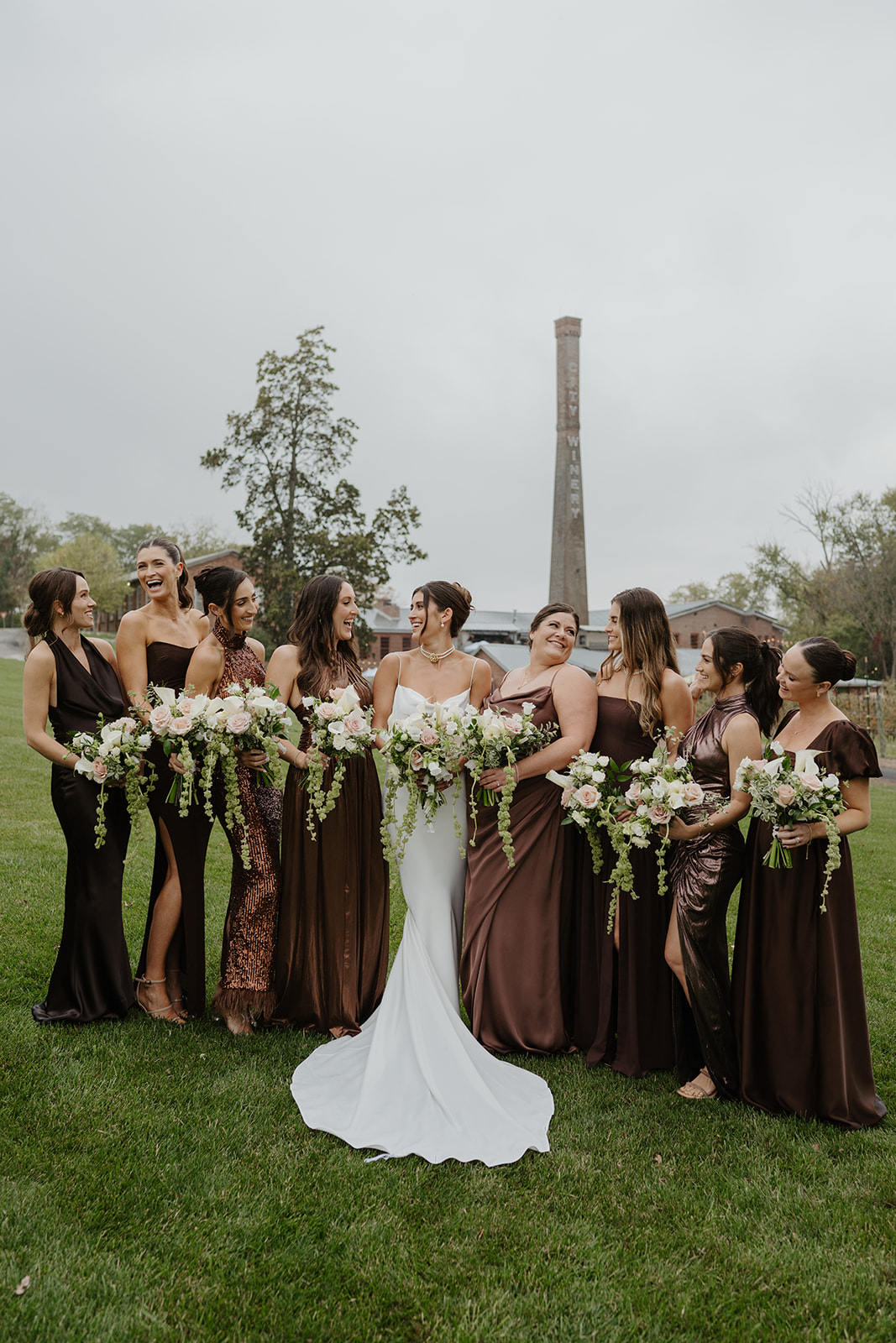 Bride smiles with her bridesmaids wearing elegant brown and bronze dresses on the lawn.