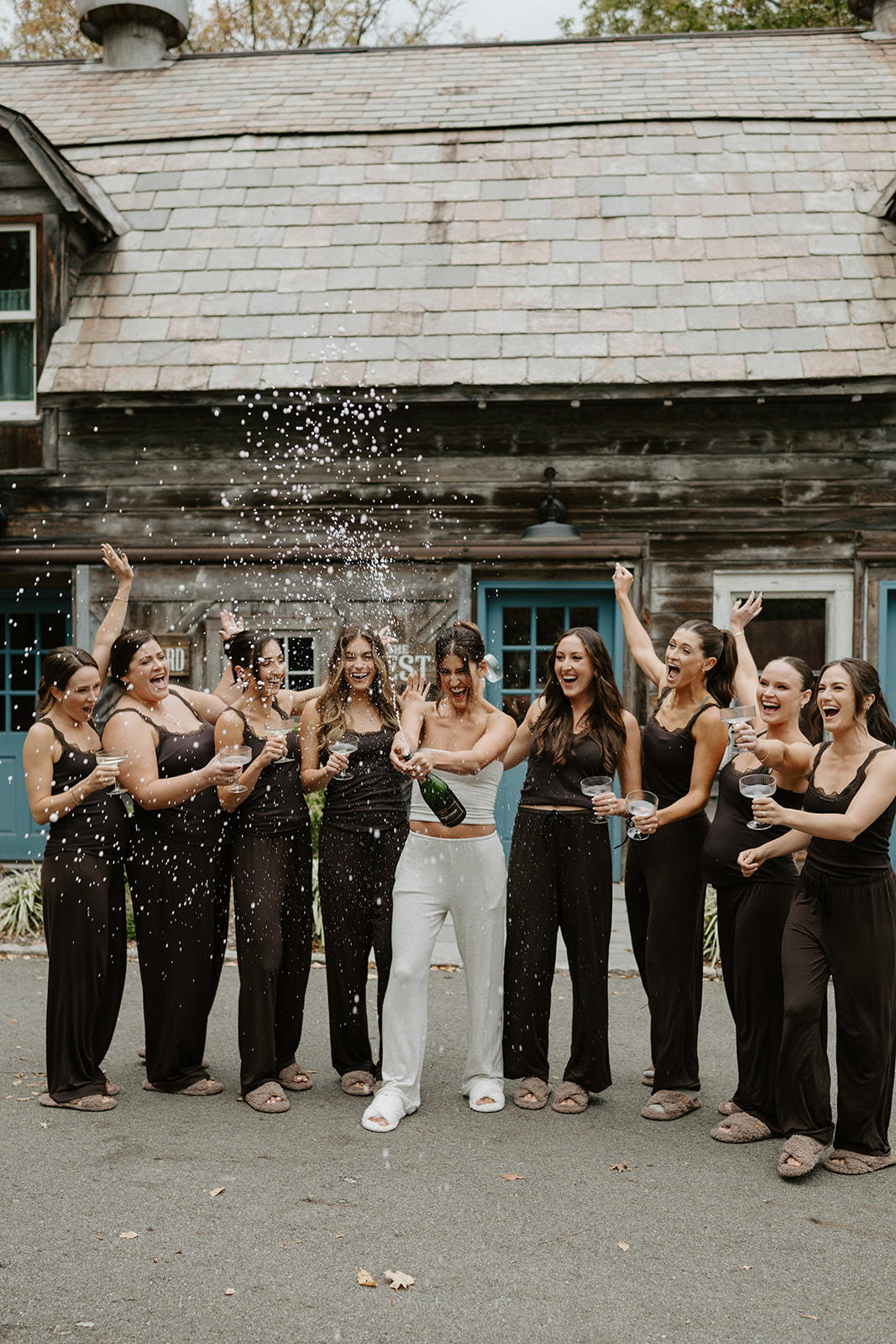 Bride sprays a bottle of champagne as bridesmaids in matching pajamas cheer outside a rustic cottage.