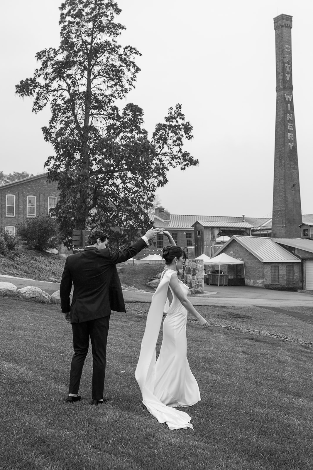 Black and white photo of the groom spinning the bride on the lawn, with the historic smokestack behind them.