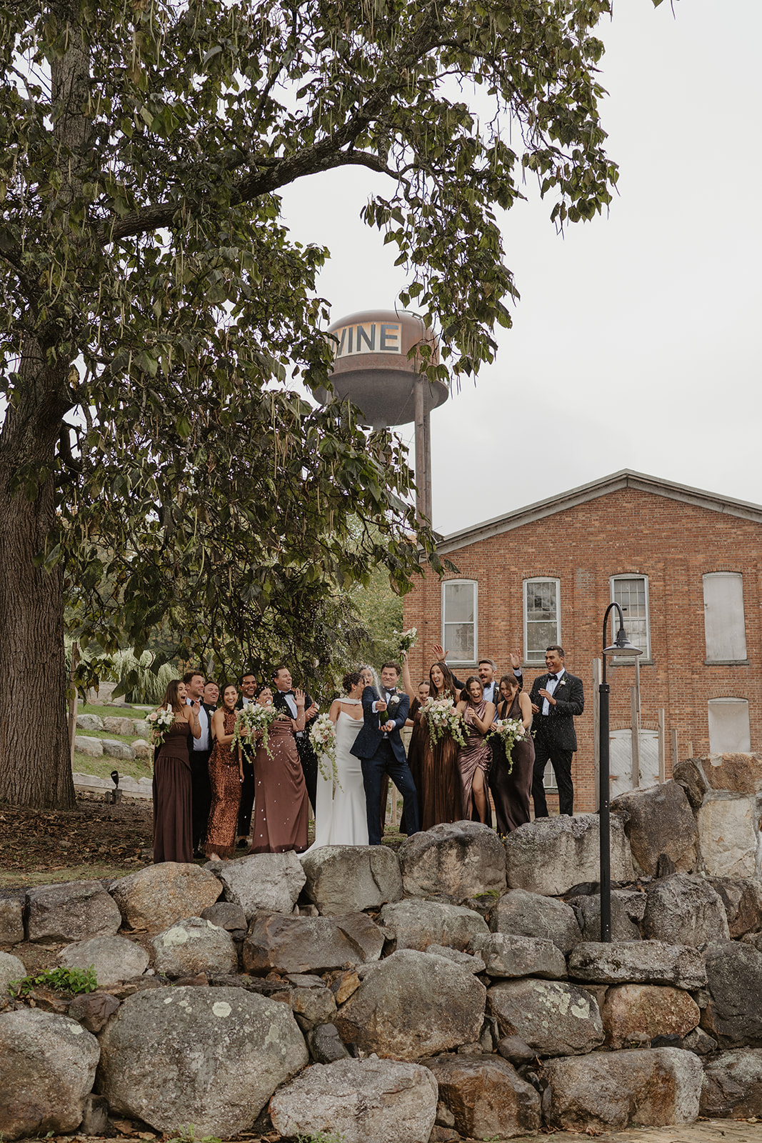 Groom pops champagne while the wedding party cheers from a stone terrace by a brick warehouse.