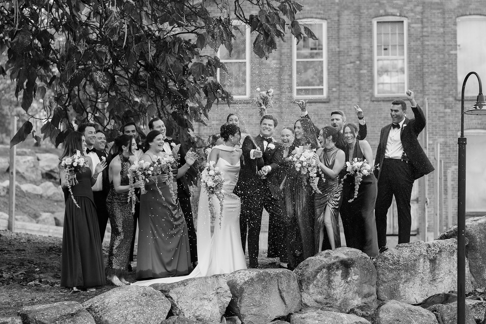 Black and white photo of the groom popping champagne as the wedding party laughs and cheers.