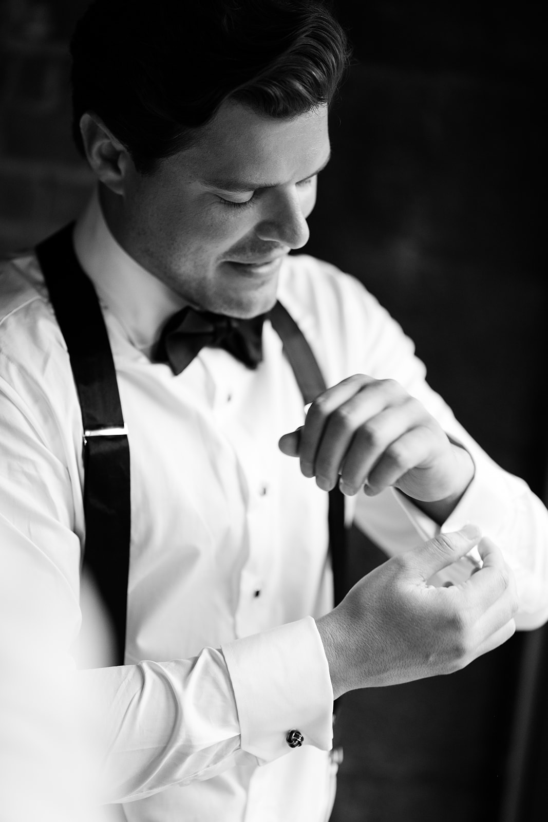 Black and white portrait of the groom adjusting his cufflinks while looking down with a soft smile.