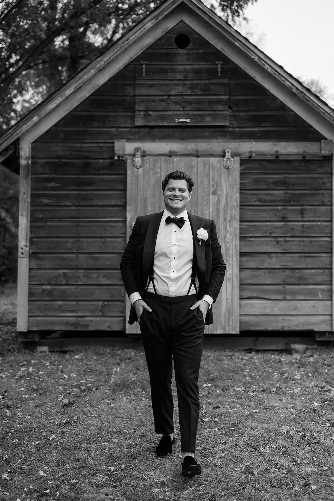 Black and white photo of the groom smiling as he walks in front of a rustic wooden shed.