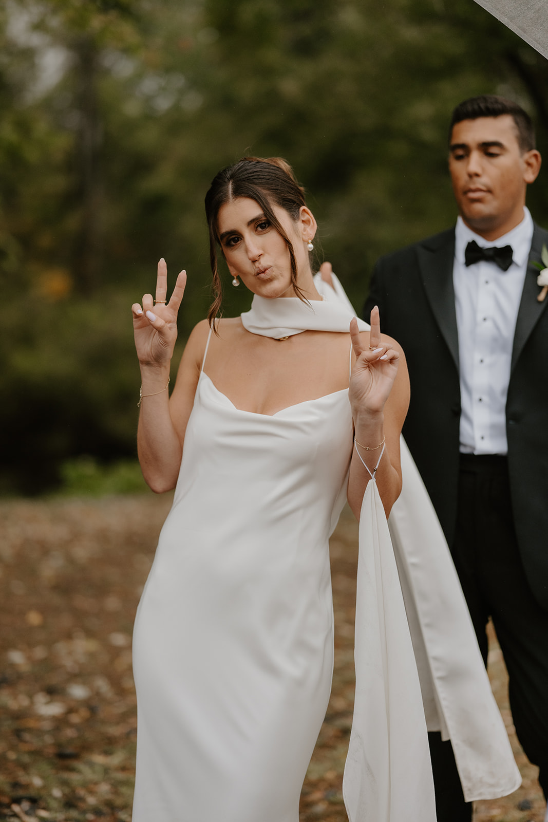 Bride flashes peace signs and makes a playful kissy face, with a groomsman blurred behind her.