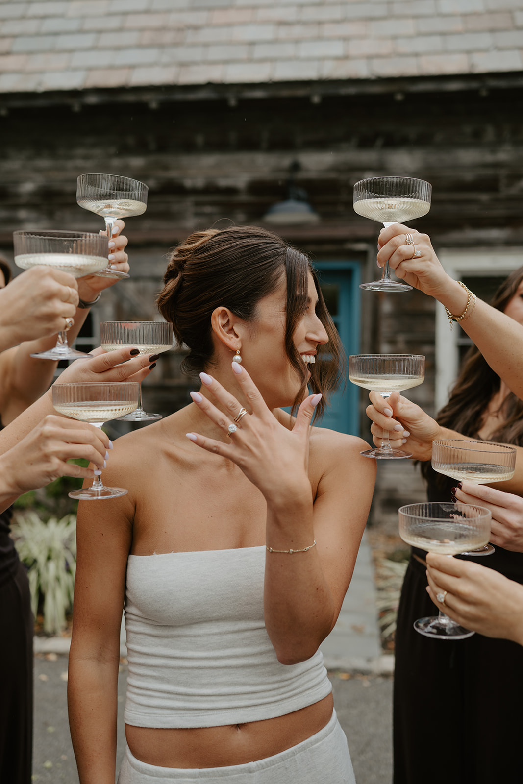 Bride admires her engagement ring as bridesmaids raise champagne coupes around her.
