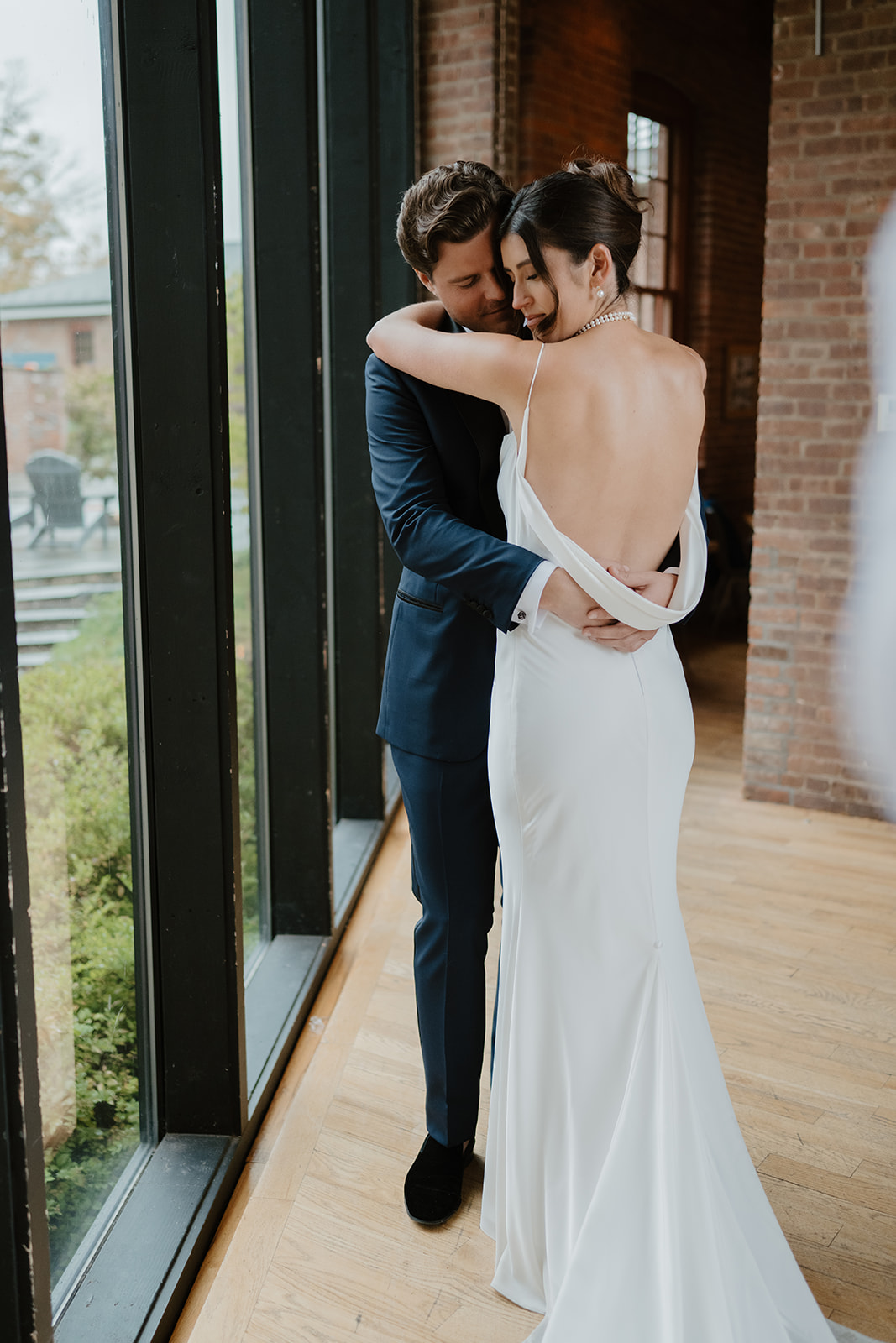 Bride and groom embrace tenderly by large floor-to-ceiling windows inside the brick winery.