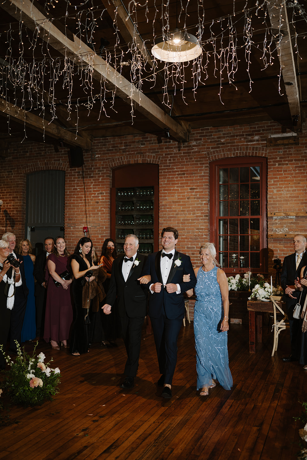 Groom walks down the aisle arm-in-arm with his parents under sparkling string lights.