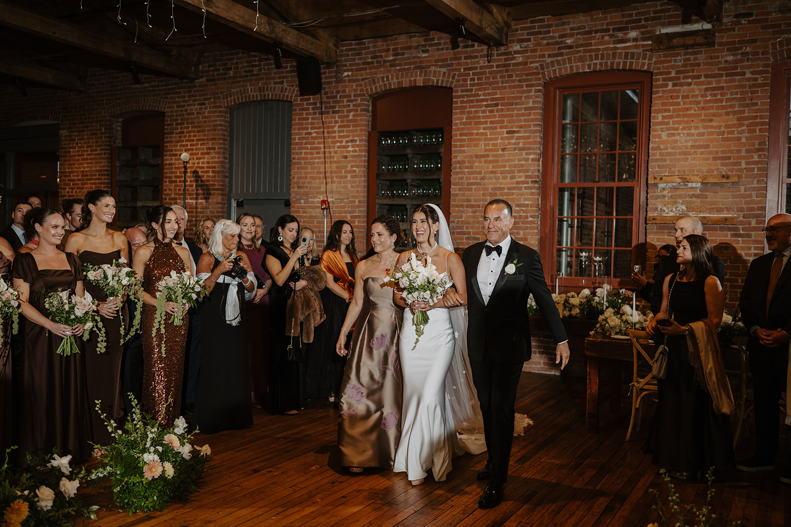 Bride walks down the aisle with both parents inside the warmly lit brick ceremony room.