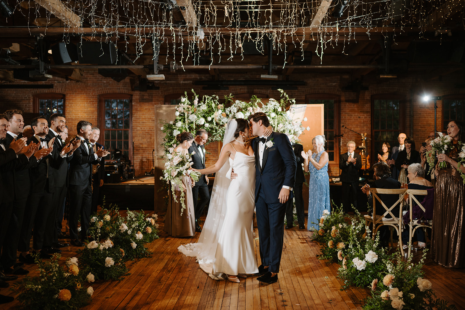Bride and groom pause for a kiss in the aisle, surrounded by clapping guests at City Winery.